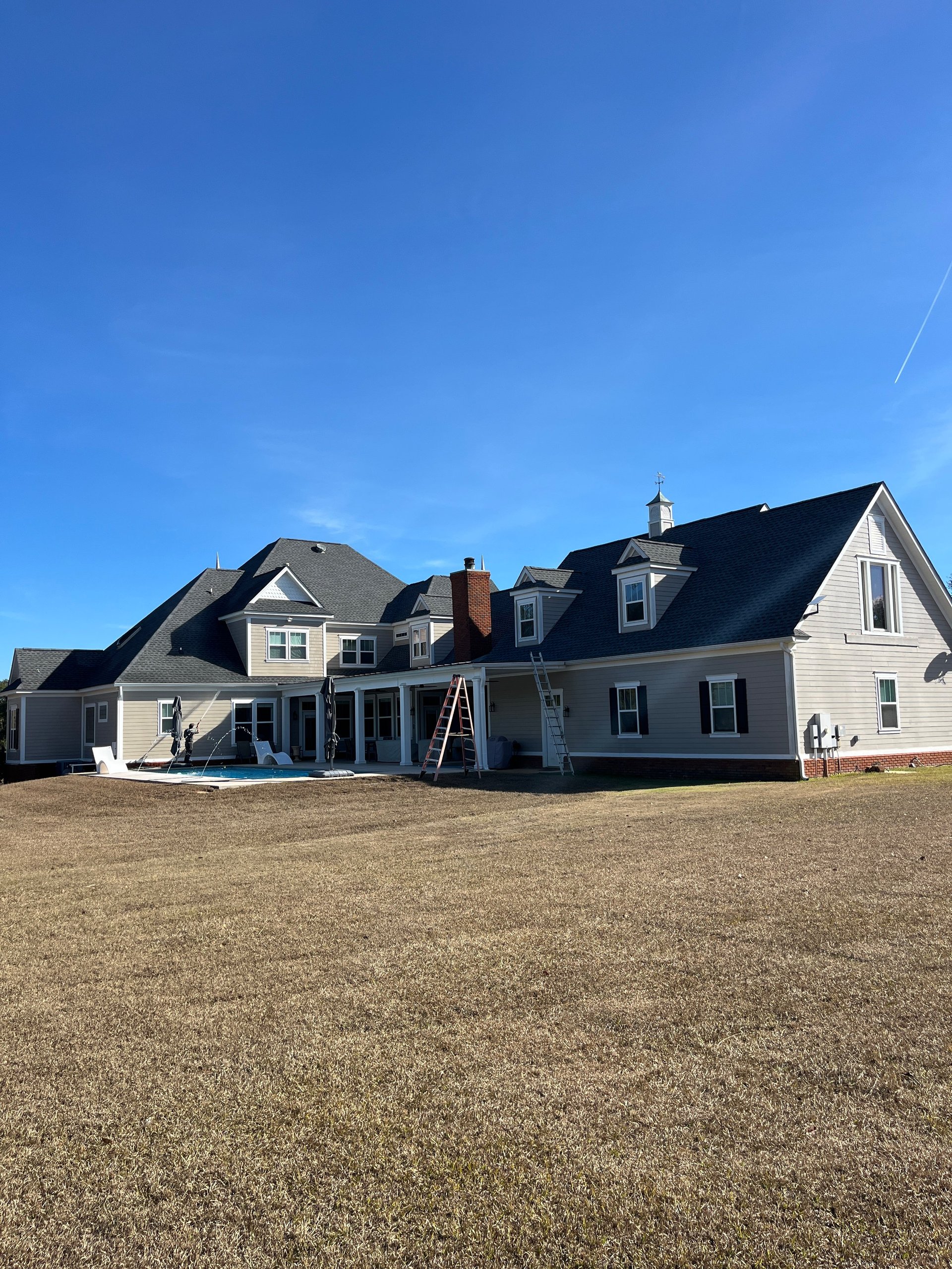 Large two-story house under construction with white siding and dark roof against a clear blue sky, set in an open grassy field