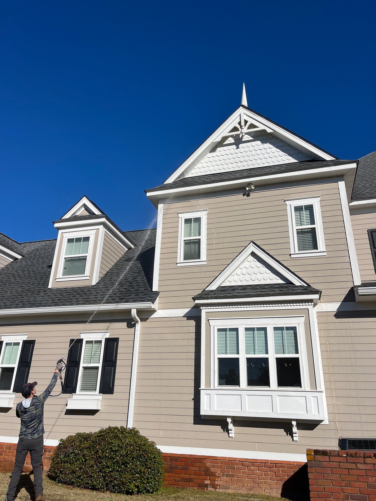 Two-story colonial-style house with beige siding, white trim, pitched roof with spires, and brick foundation under a clear blue sky